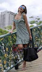 Stylish Asian woman wearing a floral dress and high-heeled sandals carries a black bag while standing on a decorative city bridge with ornate railings and lush greenery