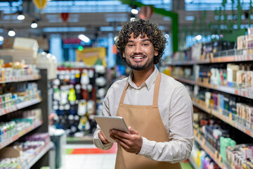 A smiling grocery store worker holds a tablet while standing in an aisle, surrounded by products on...