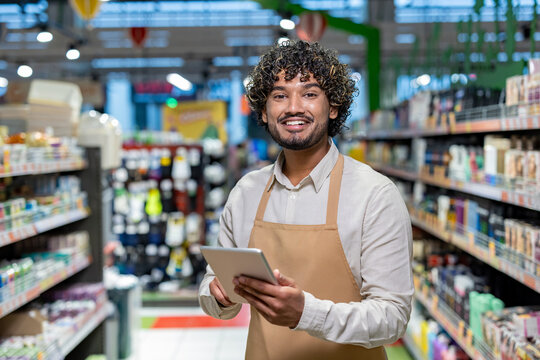 A smiling grocery store worker holds a tablet while standing in an aisle, surrounded by products on shelves.