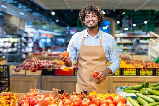 A smiling grocery store worker stands behind a counter, holding tomatoes and looking at the camera, with produce in the foreground. - Powered by Adobe