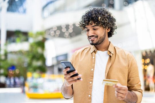 A smiling man in a shopping mall, holding a phone and a credit card, ready to make a purchase, depicting the convenience of online shopping.