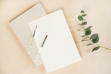 Two paintbrushes rest on a blank notepad beside textured sketch paper. A sprig of eucalyptus adds a...