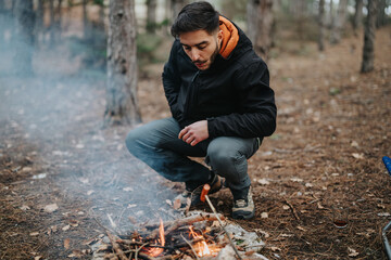 A man kneeling on the forest floor tends to a small fire, surrounded by tall trees, showcasing...