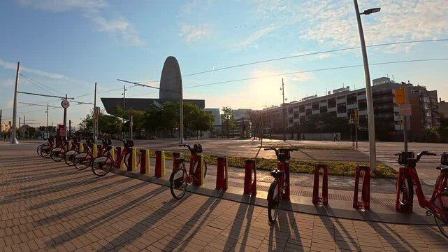 Red electric bicycles parked in a row at a bike sharing station in barcelona, spain, during sunrise, with the torre glories skyscraper in the background