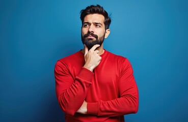 Hispanic man with beard in red sweater thinks about something. Male touches chin, looks up, isolated on blue background. Serious face expression. Studio shot, copy space for text.