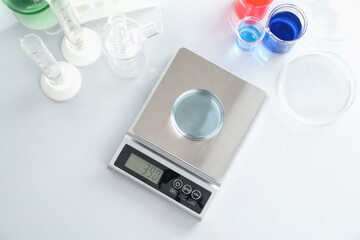 Scale with Petri dish of sample and other laboratory glassware on white table, flat lay