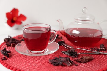 Delicious hibiscus tea in glass cup, teapot, dry roselle sepals and spoons on white table, closeup