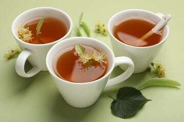 Tasty linden tea in cups, spoon, leaves and flowers on light green background, closeup