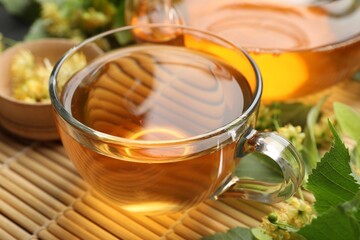 Tasty linden tea in cup, leaves and flowers on bamboo mat, closeup
