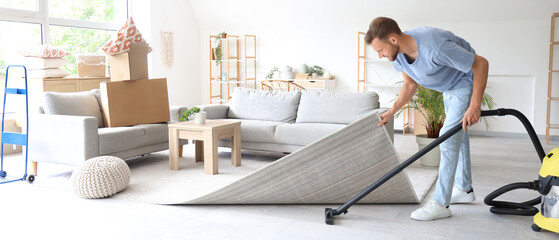 Male janitor cleaning floor under carpet with vacuum cleaner in room
