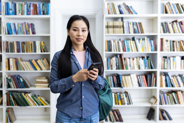 An asian woman is smiling while holding her phone in a library with many books in the background.