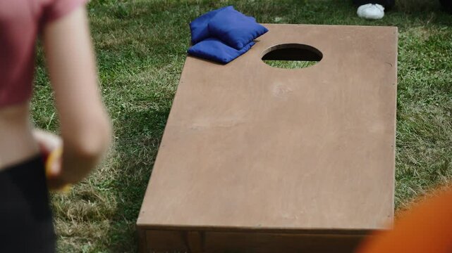 Unrecognizable female teenager throws bean bags on cornhole board. Young slim female person hits target twice with red bean bag on green lawn. Unrecognizable assistant supervises children game.