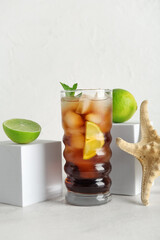 Glass of cold Cuba Libre cocktail, decorative podiums with limes and starfish on white background