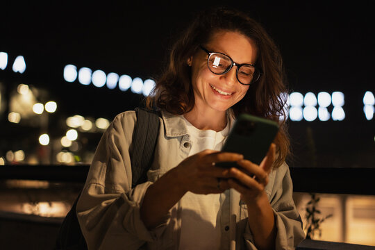 Smiling young woman uses smartphone on night city street.