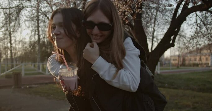 Two teenage girls enjoy a beautiful day in the park. One gives the other a piggyback ride as they walk along a path and sip milkshakes.