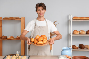 Handsome young man with board of fresh buns on table in bakery