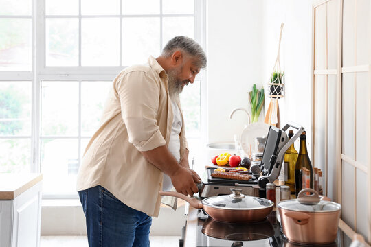 Mature man cooking tasty sausages and vegetables on modern electric grill at table in kitchen