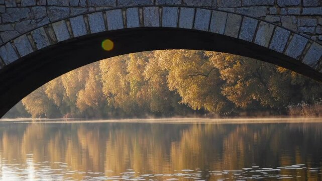 Autumn golden foliage glowing around weathered stone bridge, reflecting softly on tranquil water surface bathed in gentle morning sunlight