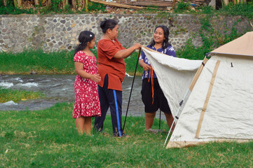 Two mothers secure the canopy guy ropes while casually chatting, as a young girl stands nearby...