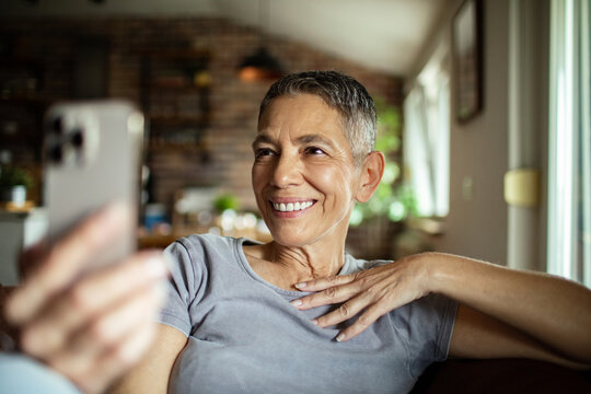 Smiling senior woman video calling family on smartphone from cozy home
