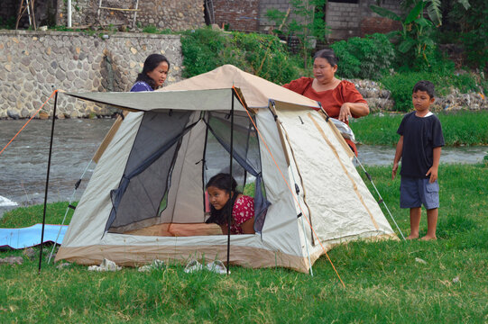 A young girl inside the tent and a boy outside observe as two mothers continue tidying the setup near the calm riverside