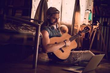 Young man playing his guitar while using a laptop in his bedroom