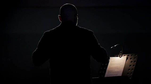 A male prompter adjusts his glasses and reads a script on a dark, backlit stage. View from behind, slow motion