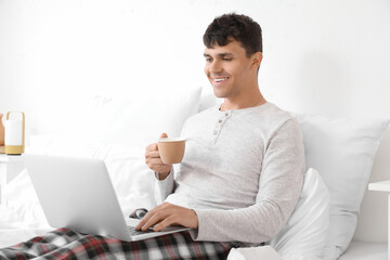 Young man with cup of coffee using laptop in bedroom
