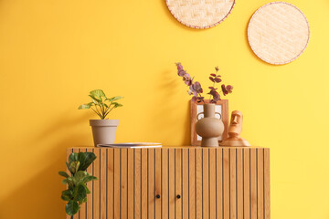 Artificial plants with decor on commode near yellow wall