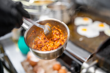 Close-up of a chef in black gloves stirring chili with beans and corn in a pot on a stove, with fried eggs cooking on a griddle in the background, in a professional kitchen setting