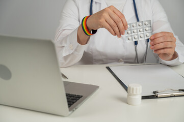 Doctor with rainbow bracelet holding pills and typing on laptop. Concept of hormone therapy for...