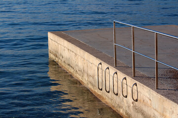 Concrete pier with metal railings and five stainless steel ladders descends into clear blue water, providing safe access for swimmers, set against a calm sea in bright sunlight