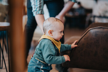 Young child holding onto furniture for support while standing, assisted by an adult in a cozy,...
