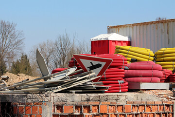 Construction site features stacks of red and yellow coiled pipes, a pile of metal road signs, a red...