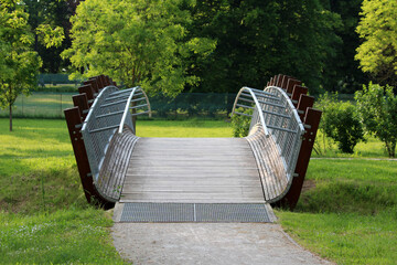 Contemporary pedestrian bridge with curved wooden deck and metal railings spans a grassy park,...