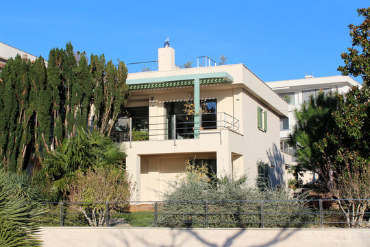 Contemporary two-story villa with flat roof, metal railings, and green pergola sits among tall cypress, palm, and pine trees, surrounded by manicured shrubs under a clear blue sky
