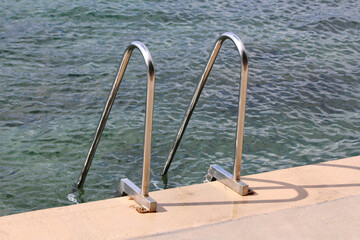 Shiny stainless steel ladder descends from a concrete edge into clear blue-green water, likely at a waterfront or swimming area, with gentle ripples and a sunlit surface © hecos
