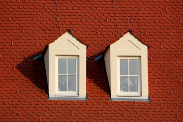 Two white dormer windows with triangular pediments stand out on a steep red tiled roof, casting sharp shadows, with windowpanes reflecting sunlight in a classic architectural style.