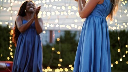 Two women clapping joyfully while wearing blue dresses outdoors   - Powered by Adobe