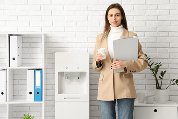 Beautiful young businesswoman with cup and folder near modern water cooler in light office