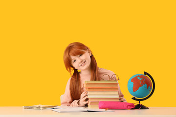 Teenage redhead girl with books at table on yellow background