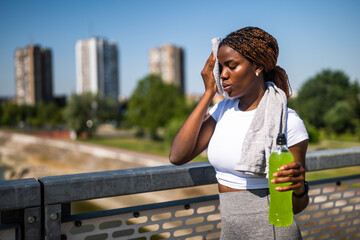 Portrait of young African-American woman in sportswear wiping sweat with towel and drinking energy...