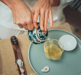 Female hands pouring tea from patterned clay teapot into clear glass pitcher, creating serene tea ritual. Soft light, wooden tea tray and greenery evoke calming mindful chenese ceremony atmosphere.