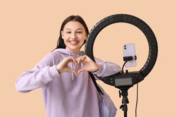 Teenage blogger with ring light making heart gesture on beige background