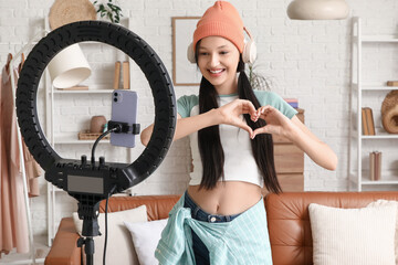Teenage blogger in headphones making heart gesture while recording video at home