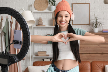 Teenage blogger in headphones making heart gesture while recording video at home