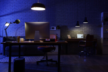 Interior of dark programmer's office with desks, sofa and glowing lamps at night