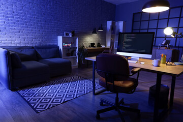Interior of dark programmer's office with desks, sofa and glowing lamps at night