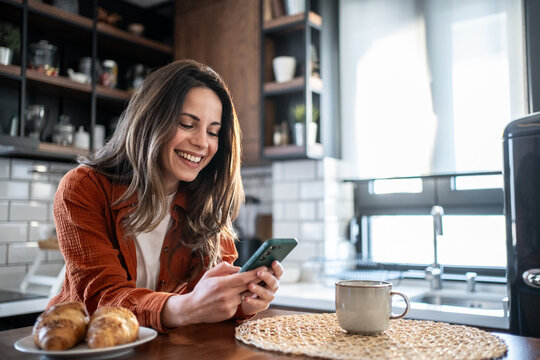 Happy woman using smartphone and having breakfast in modern kitchen