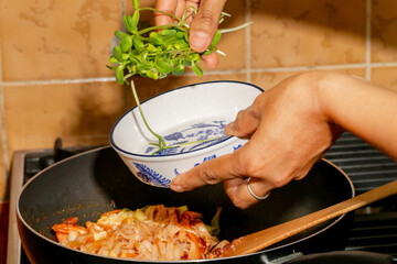 A South Asian female hand scatters vibrant microgreens into sizzling stir-fry, celebrating the Harvest Moon Festival and Earth Day